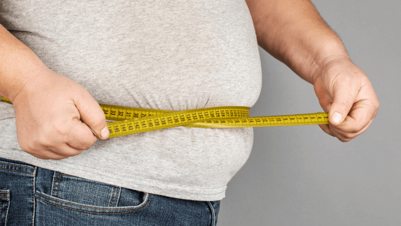 A man measures his fat belly with a measuring tape. on a gray background