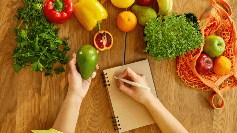 Woman planning, writing weekly meals on a meal planner note or diet plan on wooden table with healthy food fruit and vegetables in her kitchen at home.